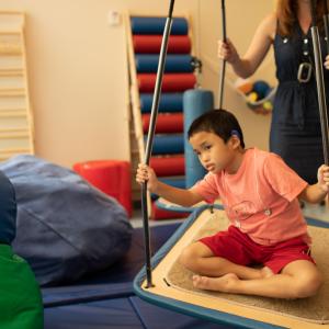 Manhattan Star Academy student is in therapy room sat criss cross on a platform swing, an adult is behind them supporting/moving the swing