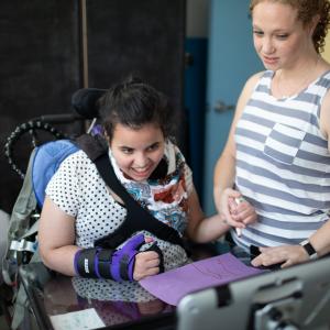 iHOPE student is in a wheelchair and looking at a tablet and is drawing on paper with support from therapist holding their hand