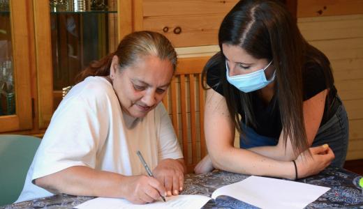 Two people sit at at table. One on the left is writing on a piece of paper, the other on the right is looking on and wears a mask.