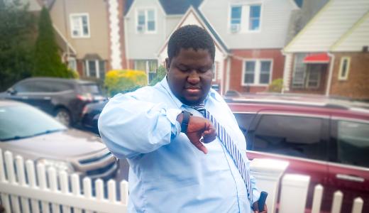 Person in blue button down shirt and tie stands outside looking at his watch. You see houses and cars in the background