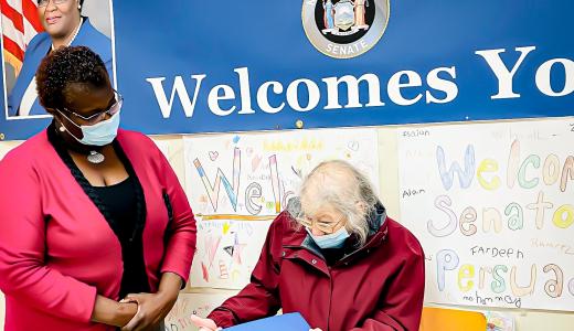 Two women stand in front of a wall with printed and hand made posters. One has a walker and holds a folder while the other looks at them. They both wear masks