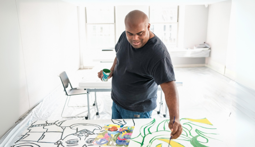 Jimmy stands in a relatively empty room with a large canvas in front of him. He holds a cup of paint in one hand and a paintbrush in the other