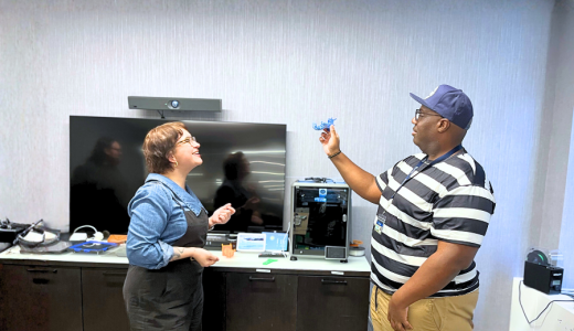 Two people stand in a room with various technological items. They are facing one another while the person on the right holds up a 3D printed item