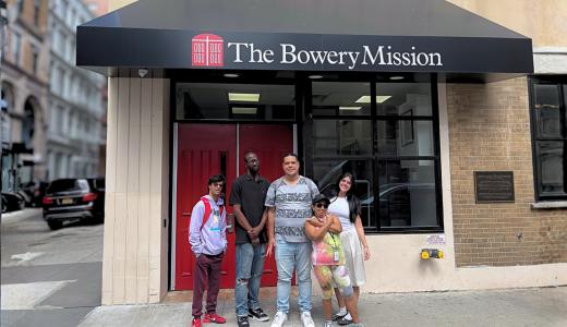 Group of people standing in front of a storefront entrance labeled “The Bowery Mission” with glass doors and brick walls on a New York City street.
