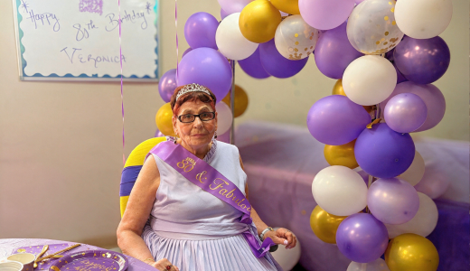 Veronica is seated indoors at her 80th birthday celebration. She is wearing a purple sash and glasses, with a large purple, white, and gold balloon cluster behind her and a hand-drawn “Happy Birthday” message on the wall.