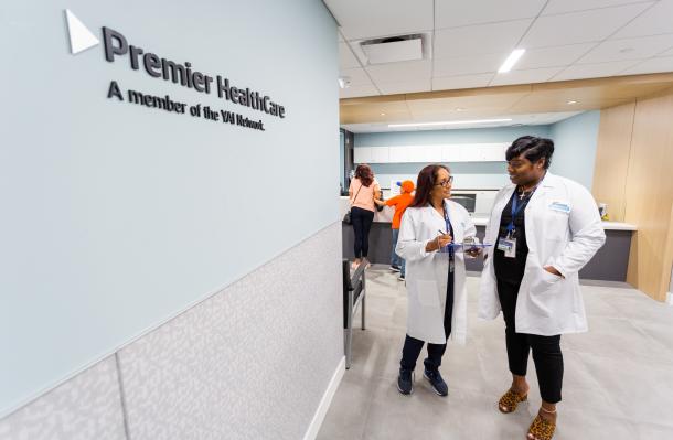 Two people wearing white coats stand in a large room/hallway with the Premier HealthCare logo on the wall to the left of them