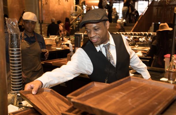Man behind the counter at a Starbucks store, he wears a flat cap and is smiling as he works