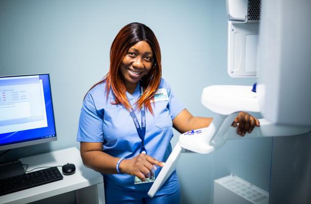 Healthcare professional stands in a dental office setting up x-ray machine. They smile at the camera and there's a computer monitor behind them on the left