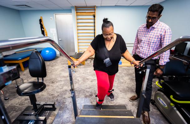 Two people stand in therapy room, person in center is holding banister and about to take a step up or down on the steps in from of them. Therapist stands alongside watching