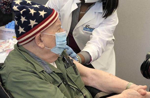 Man in profile wears a blue hat with white stars, green button down shirt, and a mask is getting a vaccination in his left arm