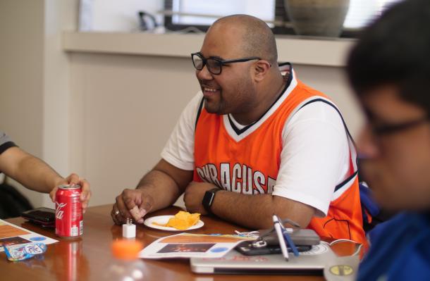 Person sits at a table with a plate of food and can of soda in front of them. They appear to smiling and talking to another person out of the shot