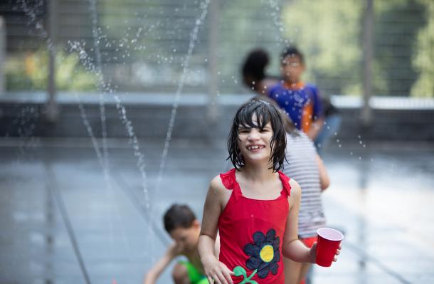 Manhattan Star Academy student outside playing in water sprinkler, they wear a red bathing suit and holding a red cup