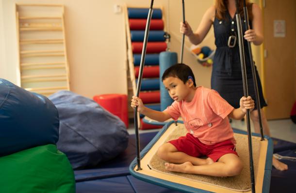 Manhattan Star Academy student is in therapy room sat criss cross on a platform swing, an adult is behind them supporting/moving the swing