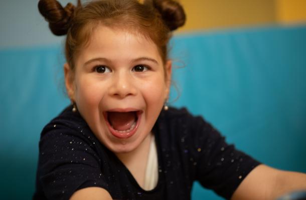 Young child with hair in 2 buns wearing a black top has a big, excited smile, the background is teal