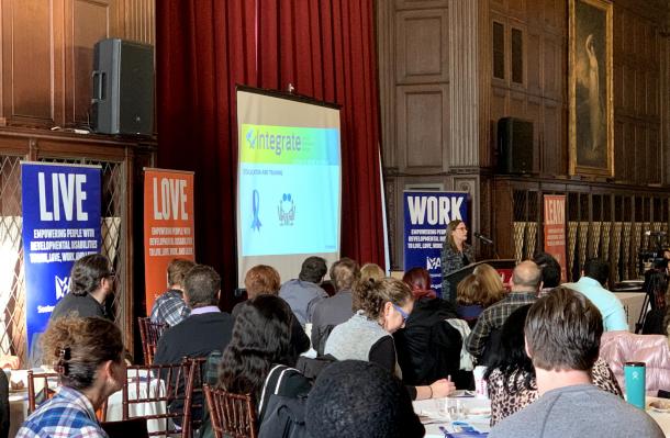 Group of people sat around tables in a large room. In background you can see a person is standing at a podium talking, there is a slideshow behind them and conference posters around.