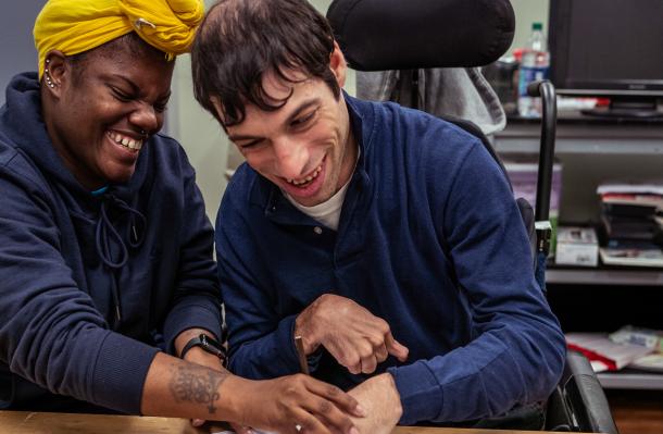 2 people sit at at table, one is in a wheelchair, they are both engaged in a drawing task with the person on the left supporting the one on the right. they are both fully enjoying what they are doing with big smiles