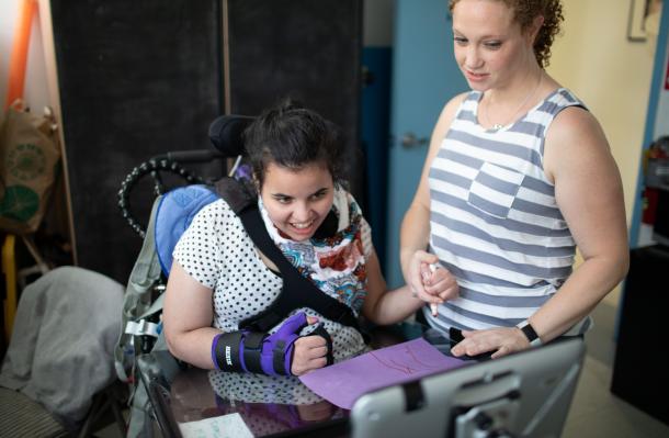 iHOPE student is in a wheelchair and looking at a tablet/communication device with support from therapist holding their hand