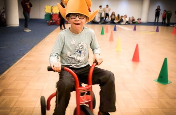 MSA student on a tricyle wearing a turkey hat and glasses. They are looking at the camera and you can see they are in a gym with people and colorful cones in the background