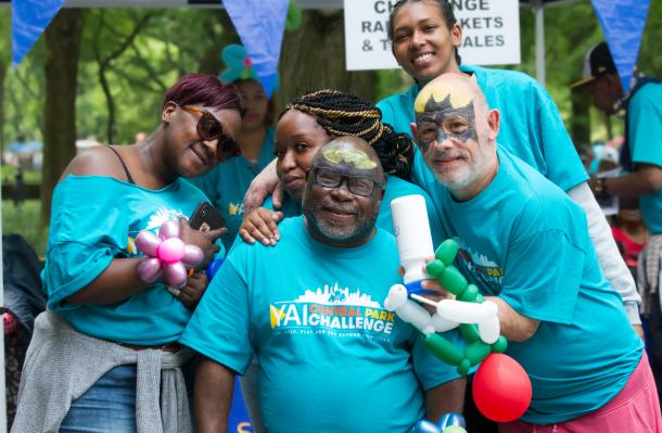 Group of 5 people wearing teal Central Park Challenge shirts pose for photo in central park. The 2 in the forefront have on batman facepaint