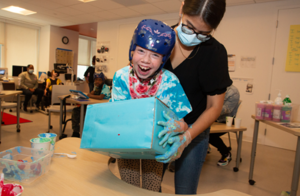 iHOPE student supported by staff while they hold an aqua colored box, they both have aqua colored hands and the student has a great big smile