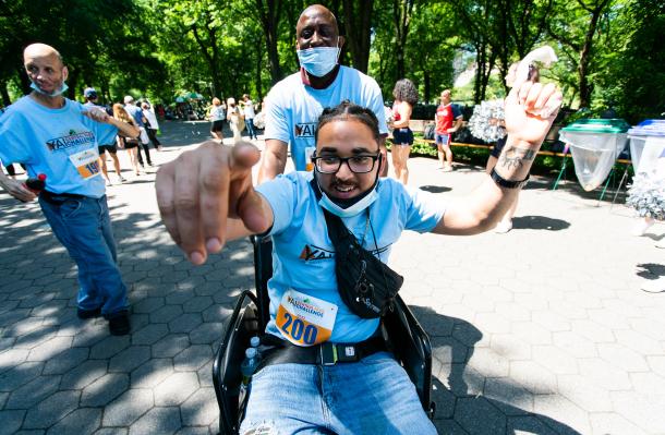 Young person in a wheelchair points at camera with left arm up. Someone else stands behind them. There are people all around in in the background in Central Park