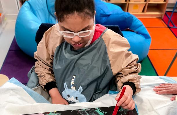 iHOPE student wearing glasses is sitting at table paining with some bright yellow paint.