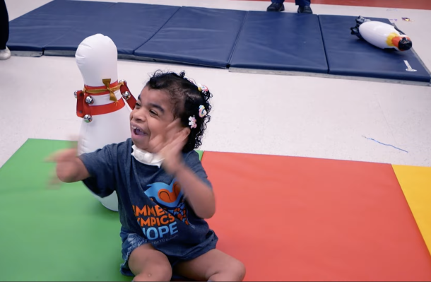 Thumbnail from video, iHOPE student is on a colorful gym mat and waving their hands and smiling