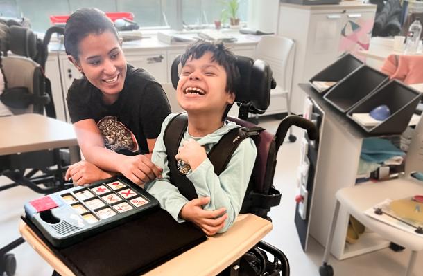 An iHOPE student and staff are sharing a laugh together, the student has a communication device in front of them on the wheelchair table