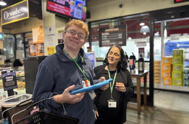 Connor stands in a supermarket with a staff member from MSA, he's holding a tablet that helps him with his shopping