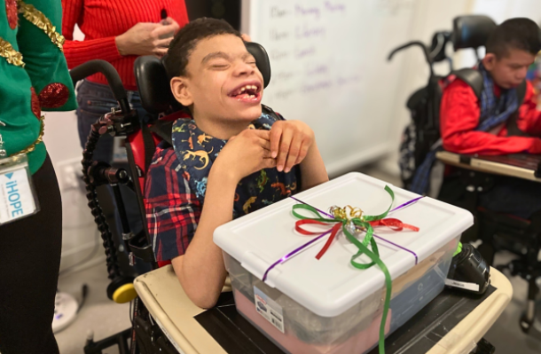 Student smiles a big smile, in front of them on the wheelchair table is a plastic box with colorful ribbon