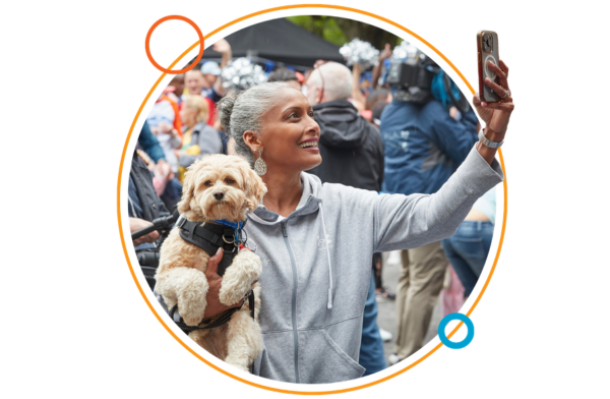 Woman in central park holds dog in one arm and her phone up in the other hand