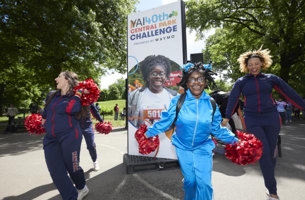 A person supported by YAI is holding red pom poms and dancing with NY Cheer cheerleaders in front of a CPC sign with their picture.