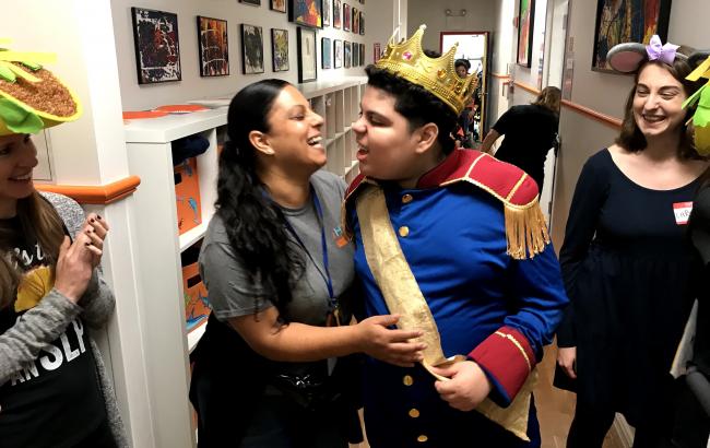 Evelyn and Ethan stand close to one another in a hallway, Ethan is smiling intensely while other people look on smiling