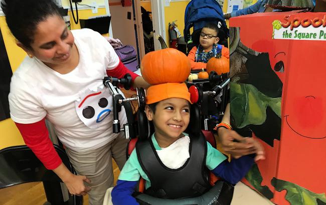 Jabir is in a wheelchair wearing an orange hat. Anitha is stood behind looking on.