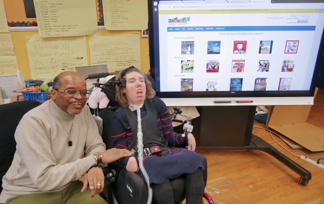 Jerry sits next to Catie, they are to the left of the image, behind them to the right is a smartscreen with icons on it.