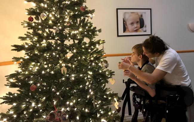 A profile view, Katherine stands behind Nick who is using a walker, they are to the right of the image with a Christmas tree to the left.