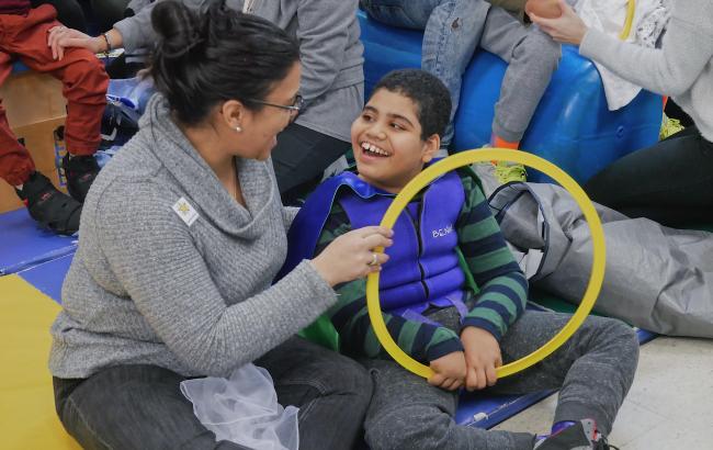 Mohammed sits on a floor mat holding a large yellow ring, he has a very big smile on his face as he looks at Andrea who is sat to his right