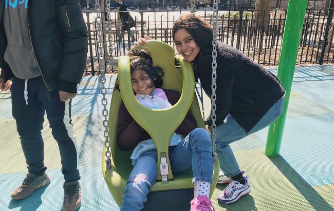 Yarisa is sat in an adaptive swing at a playground, Nicole stood beside her crouches to take a picture together. They they are both smiling.