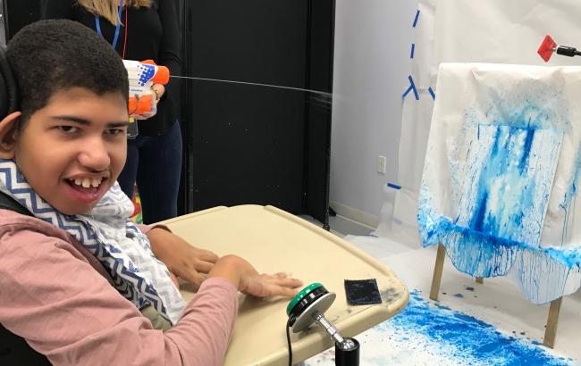 Omar had his hands on the tray of his wheelchair, in the background to the right you see paper with blue paint on it, someone in the background on the left is holding a water gun