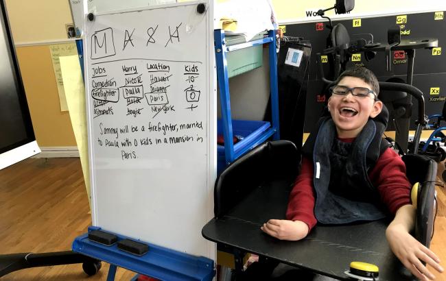 Sammy is sat in a wheelchair on the right of the picture and on the left is a whiteboard with a lot of writing at the bottom it says "Sammy will be a fire fighter, married to Paula with zero kids in a mansion in Paris"