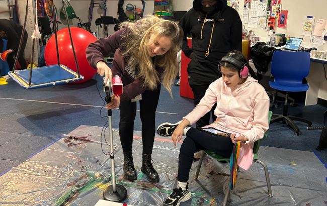 MSA student sits and looks on as a staff person leans over a canvas and looks to be pouring red paint