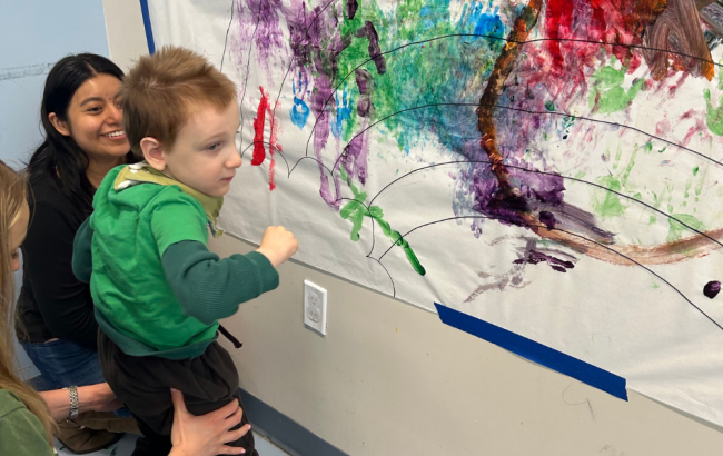 MSA student stands with support as they look on at a a colorful painting on a large sheet of paper on the wall