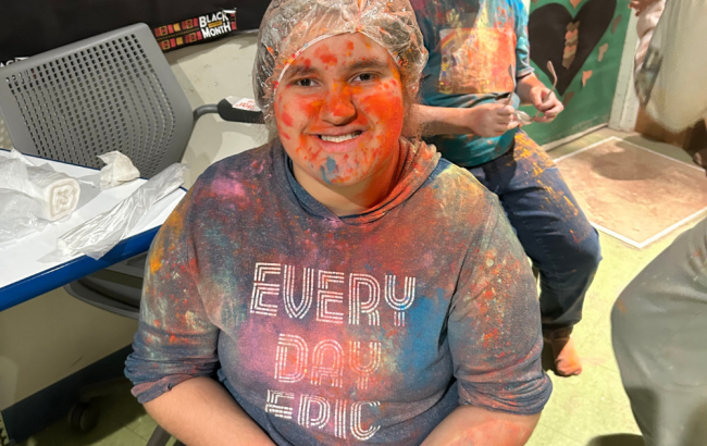 MSA student poses for camera, they are wearing a plastic cap and are covered in colorful powder