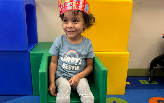 MSA student sits on a green plastic chair wearing a colorful cardboard crown