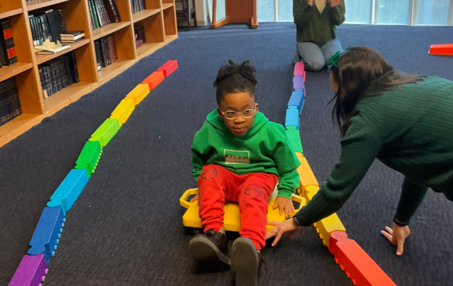 MSA student sits on a wheeled toy and is being pulled along by a staff person