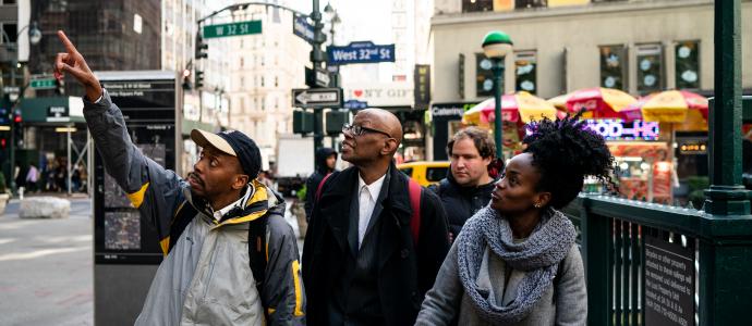 A group of people walking in bustling midtown Manhattan, one on the left is point up to the left while they all look up in that direction