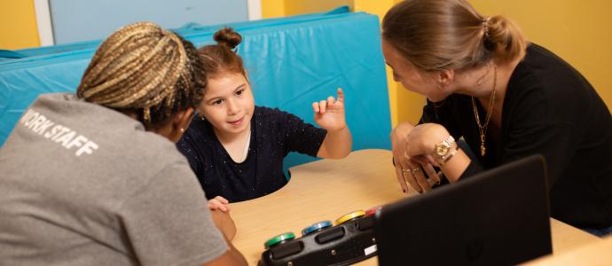 iHOPE student sits at a table and looks to be about to touch a communication device. Two staff are engaged with her and sit on either side of the device