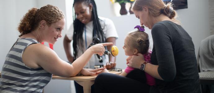 iHOPE student being supported by school staff as the student and two staff site at a school desk. A third staff person is stood in the background and appears to be engaged with another student