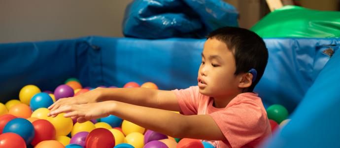 Manhattan Star Academy student is in a ball pit, their arms are outstretched, they wear a peach color shirt and hearing aid