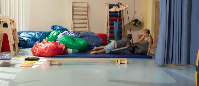 Long shot of a Manhattan Star Academy Student lays face down on a platform swing in therapy room. An adult is to the right and colorful beanbags are on the left